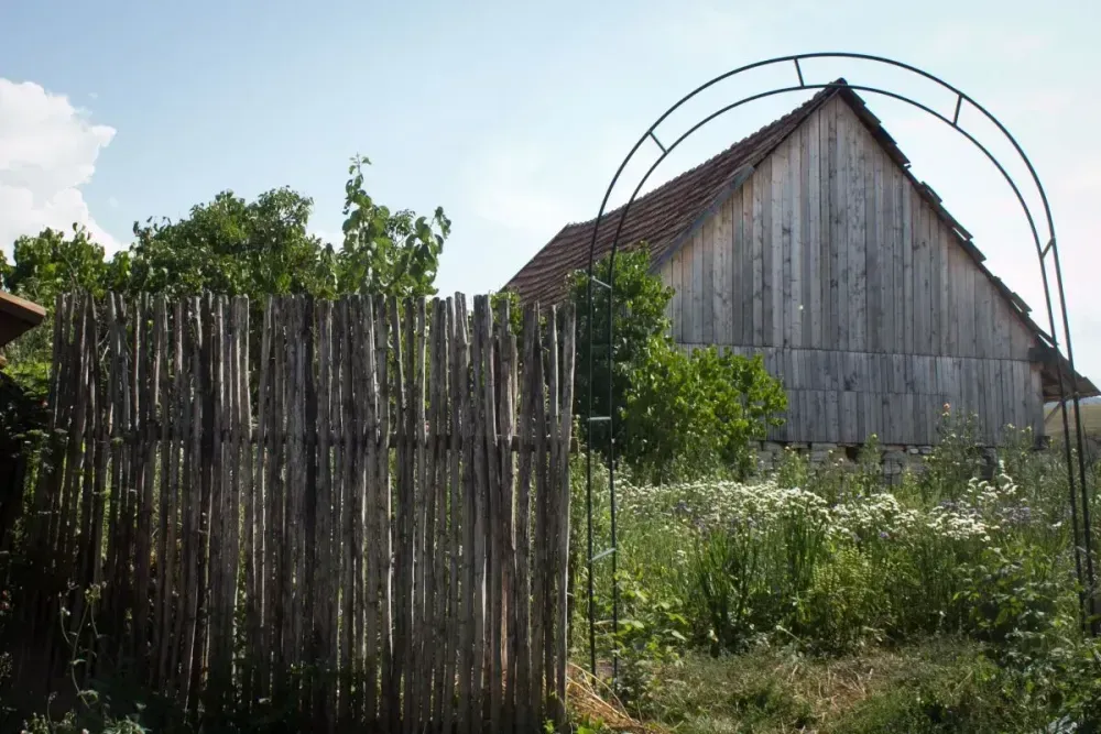 Un scenariu rural cu un gard de lemn în prim-plan, un arc metalic în spatele acestuia și o clădire tradițională din lemn sau un hambar în fundal, sub un cer senin.