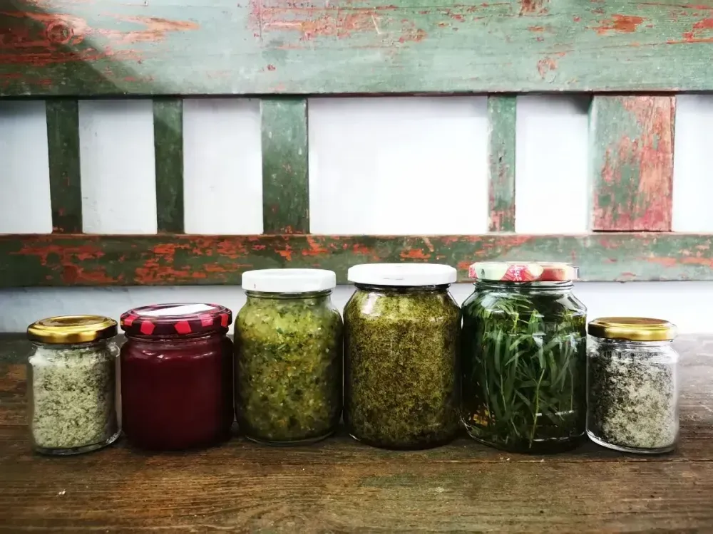 Különböző befőttesüvegek savanyított zöldségekkel és fűszernövényekkel egy rusztikus, hámló festésű fapadon.