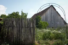 A rural scenario with a wooden fence in the foreground, a metal arch behind it and a traditional wooden building or barn in the background under a clear sky.
