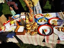 Ein Tisch im Freien mit verschiedenen Apfeldesserts, umgeben von Menschen. Im Zentrum steht ein Karaffenhalter mit Äpfeln, flankiert von Tellern mit Apfelkuchen und Apfelrosen.