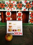 Ein Kalender für das Jahr 2024 steht vor einer Wand mit traditionellen Blumenmalereien und ein einzelner Apfel liegt im Vordergrund.