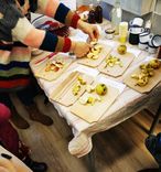 Oameni care decojesc și taie mere la o masă cu față de masă, cu pahare și sticle în fundal.